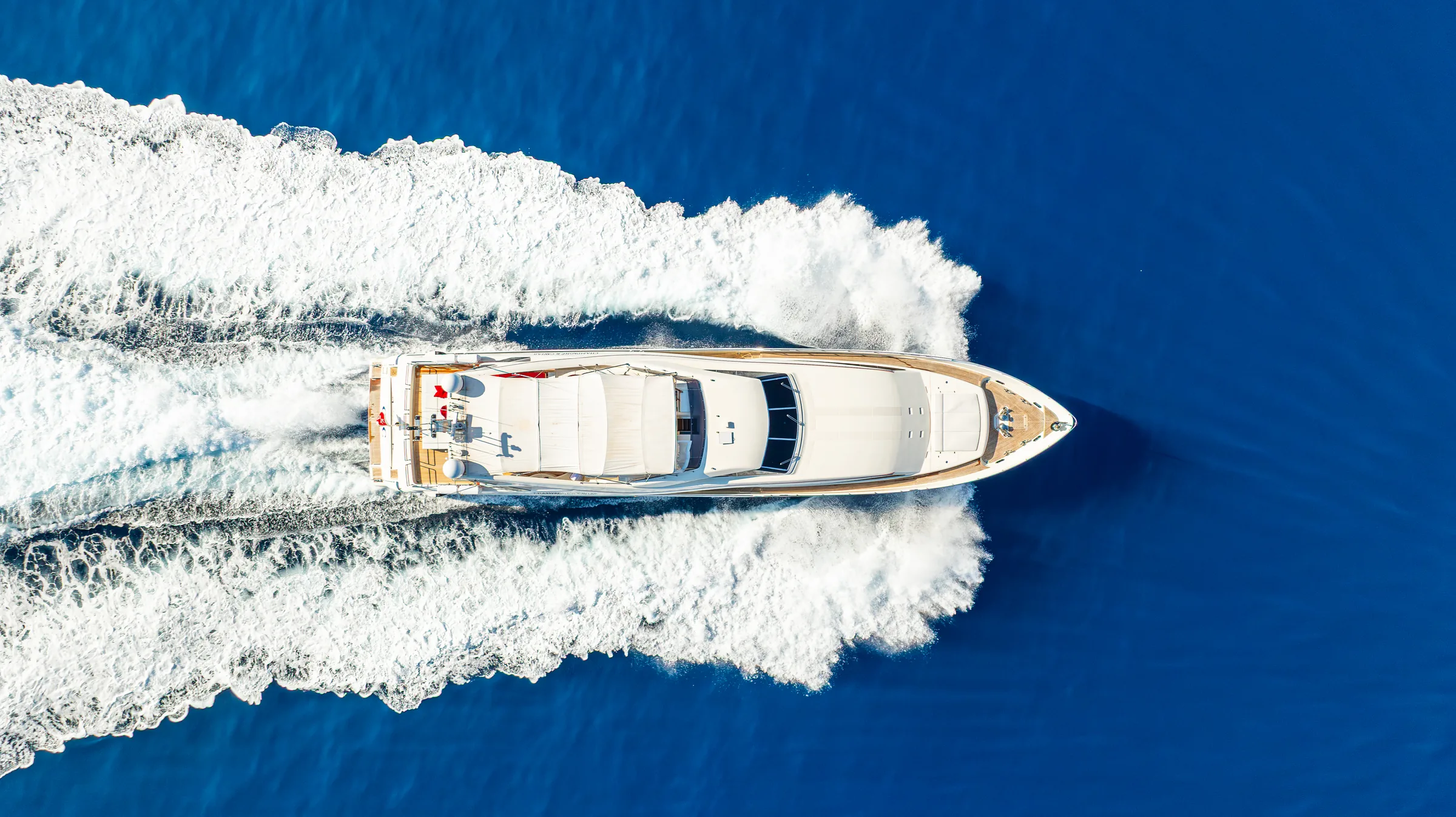 A motor yacht under way from directly above, its wake opening white across deep blue water.