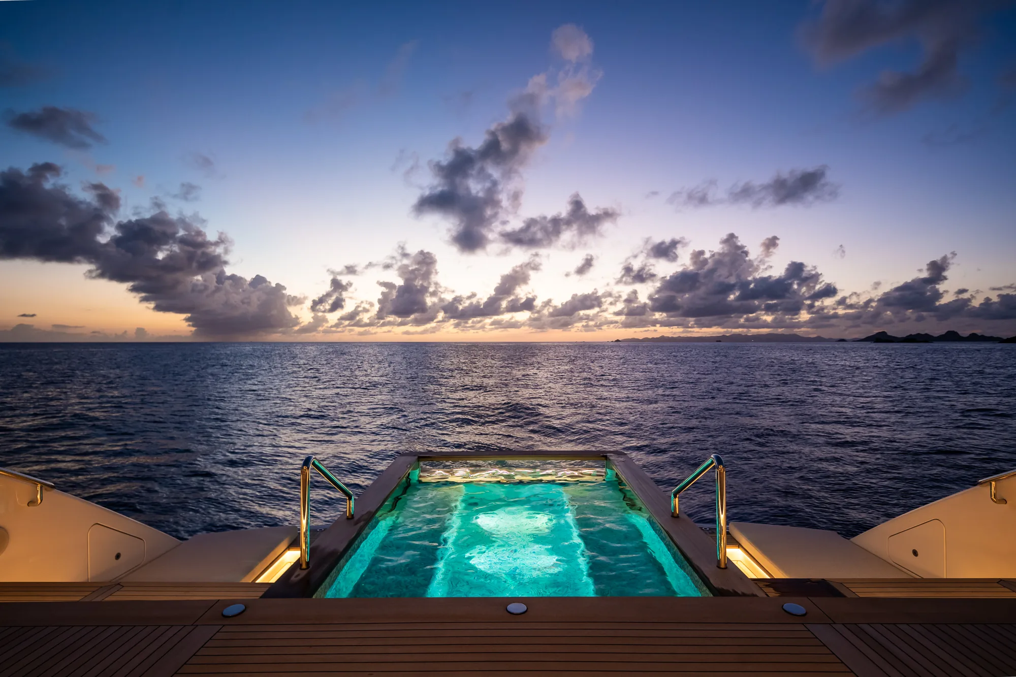 A lit deck pool on a yacht at dusk, the open sea and a broken sky beyond it.